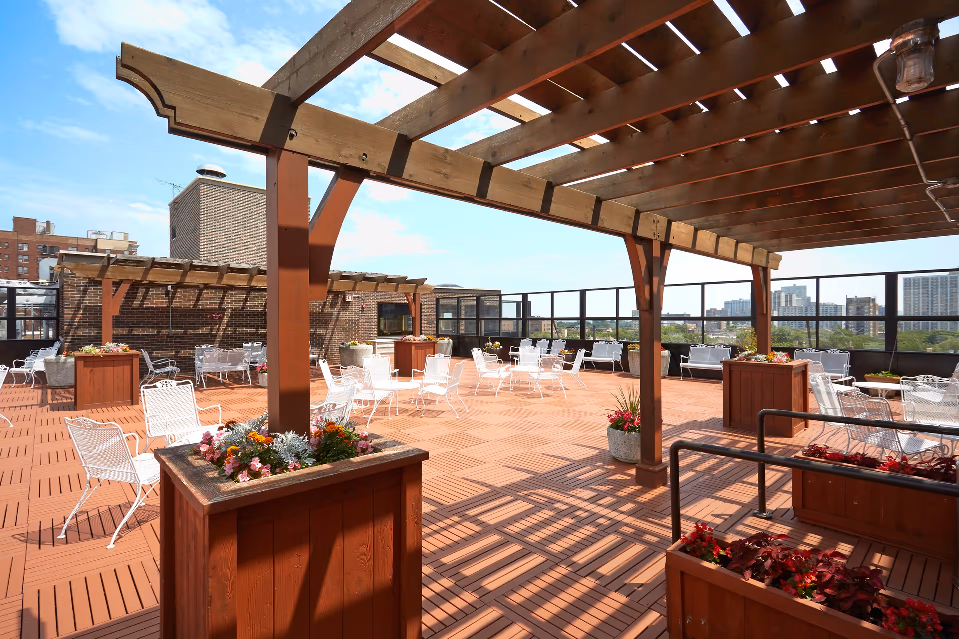 A spacious rooftop patio with wooden pergolas, multiple white metal chairs and tables, and large wooden planters filled with colorful flowers. The patio overlooks a cityscape with buildings under a partly cloudy blue sky.