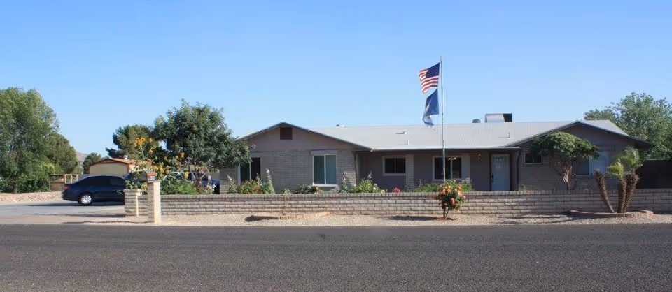 Single-story building with a gray roof and brick exterior, surrounded by a low brick wall and landscaped with small trees and bushes. Two flags, including an American flag, are flying on a flagpole in front of the building. A black car is parked on the left side of the building under a clear blue sky.