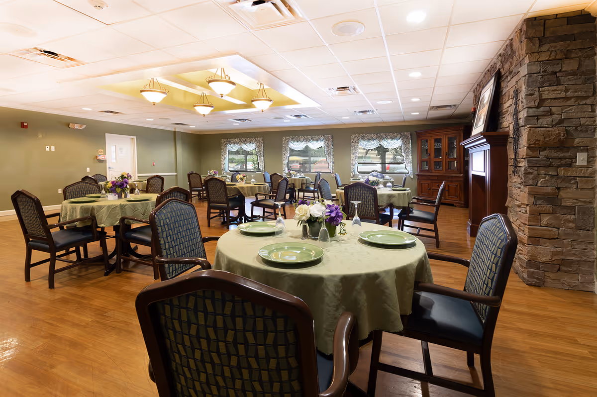 A dining room with round tables covered in green tablecloths, each set with plates, glasses, and floral centerpieces. The room has wooden floors, green walls, multiple windows with valances, and a stone fireplace on the right side. Several chairs with patterned upholstery surround the tables.