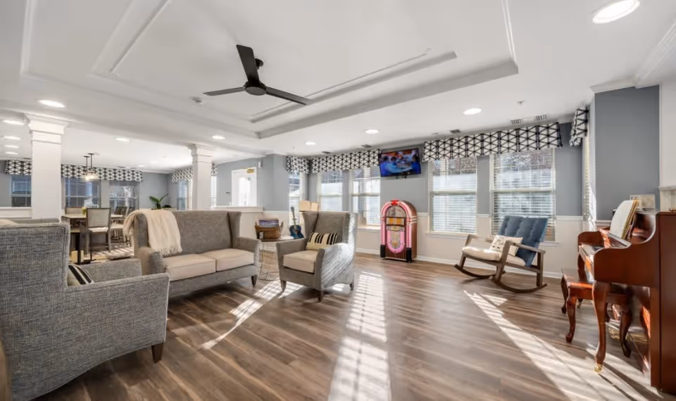 A bright and spacious living room area with large windows letting in natural light. The room features a gray sofa and matching armchairs, a wooden rocking chair, a vintage-style jukebox, and a wooden piano with a bench. The walls are painted light blue with white trim, and the floor is a medium-tone wood. There is a ceiling fan and recessed lighting, with patterned valances on the windows.