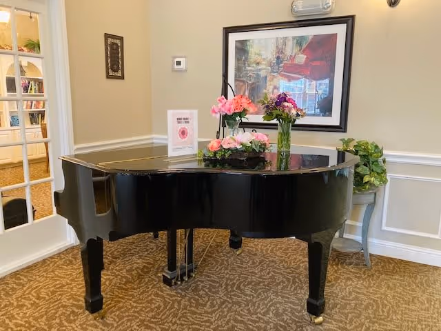 A black grand piano in a room with beige walls and patterned carpet. On top of the piano are two vases with pink and purple flowers and a small sign. Behind the piano is a framed painting of a piano and a plant on a small table to the right. A glass-paneled door is visible on the left side of the image.