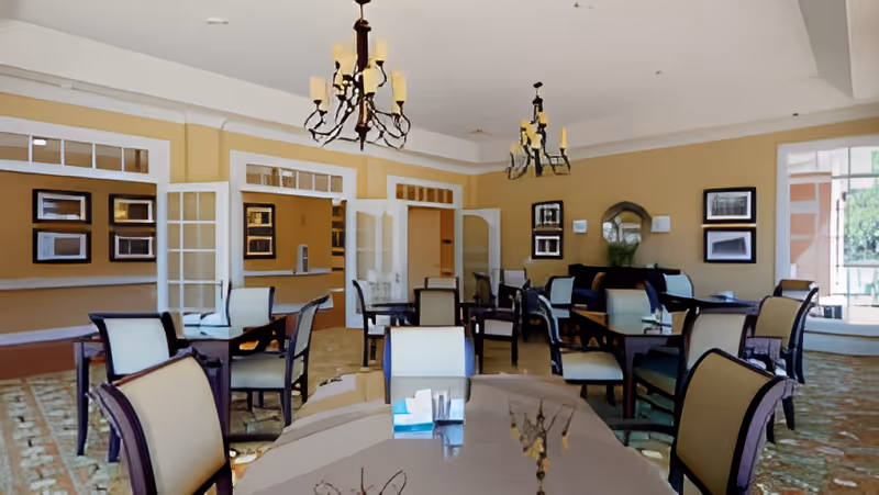 A spacious dining room with multiple tables and chairs arranged neatly. The room features beige walls, large windows allowing natural light, framed pictures on the walls, and two chandeliers hanging from the ceiling. There is a carpeted floor and glass-paned double doors leading to another area.