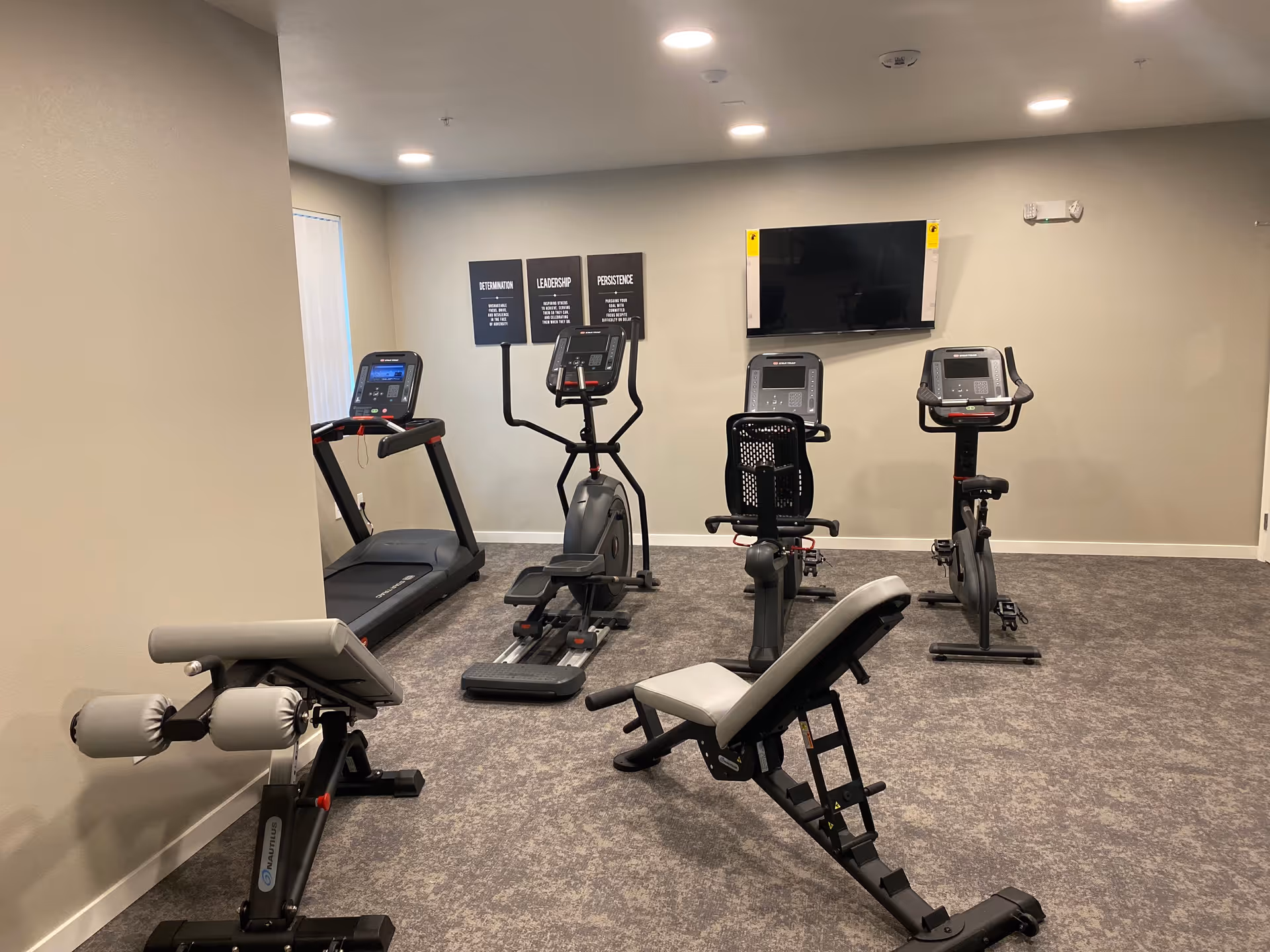 Fitness room with a treadmill, elliptical, stationary bikes, adjustable bench and wall-mounted TV.