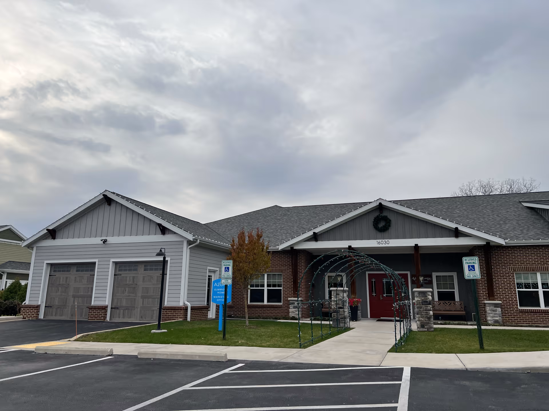Exterior front view of Azura Memory Care and Assisted Living at Mierow Farm - Elmbrook, showing a single-story building with a gray roof, brick and gray siding, a red front door, a small lawn with a tree, and a parking lot with reserved handicap parking spaces.