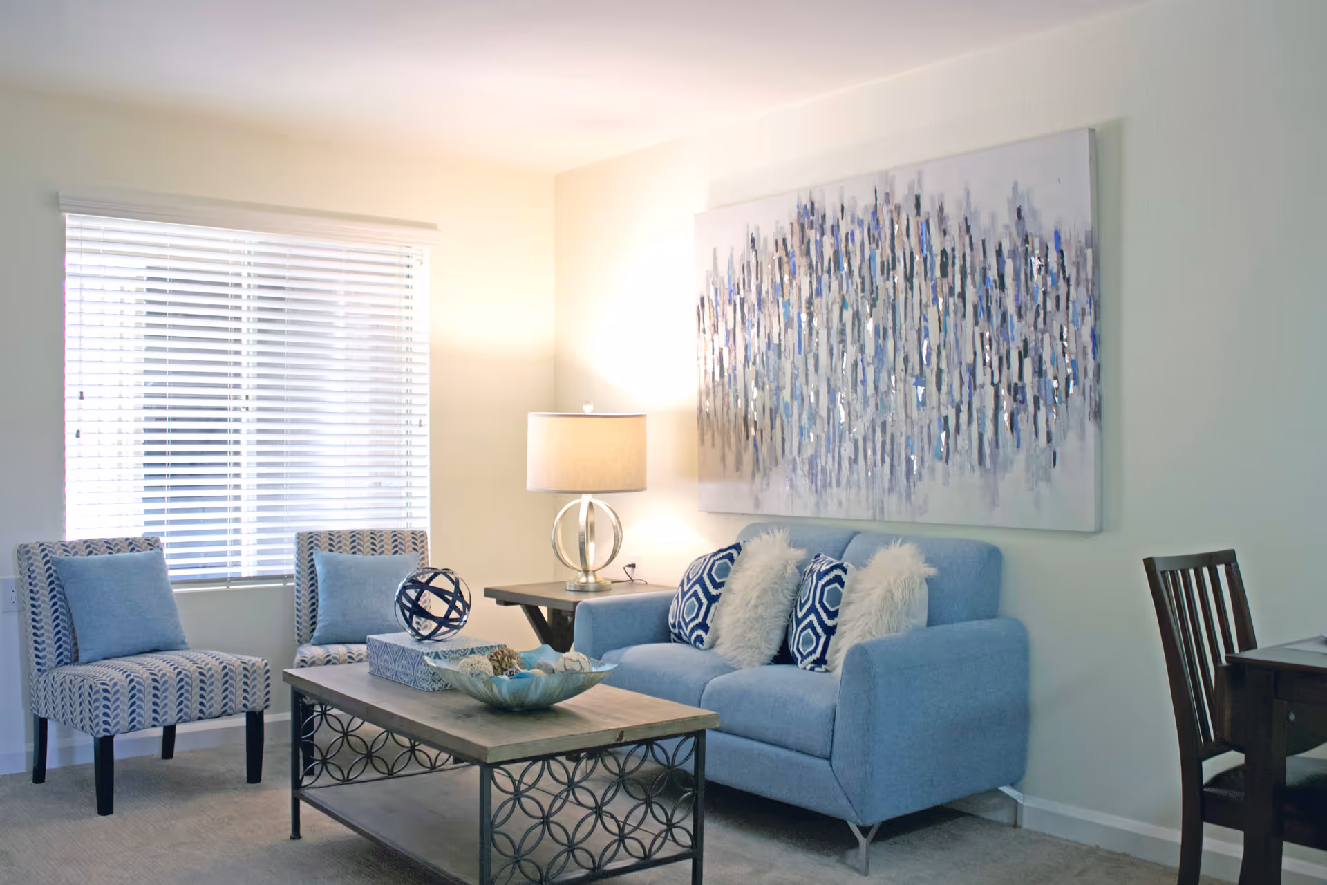 A bright living room with a light blue sofa adorned with patterned and fluffy pillows, two patterned armchairs with blue cushions, a wooden coffee table with decorative items, a side table with a modern lamp, and a large abstract painting on the wall. A window with white blinds allows natural light into the room.