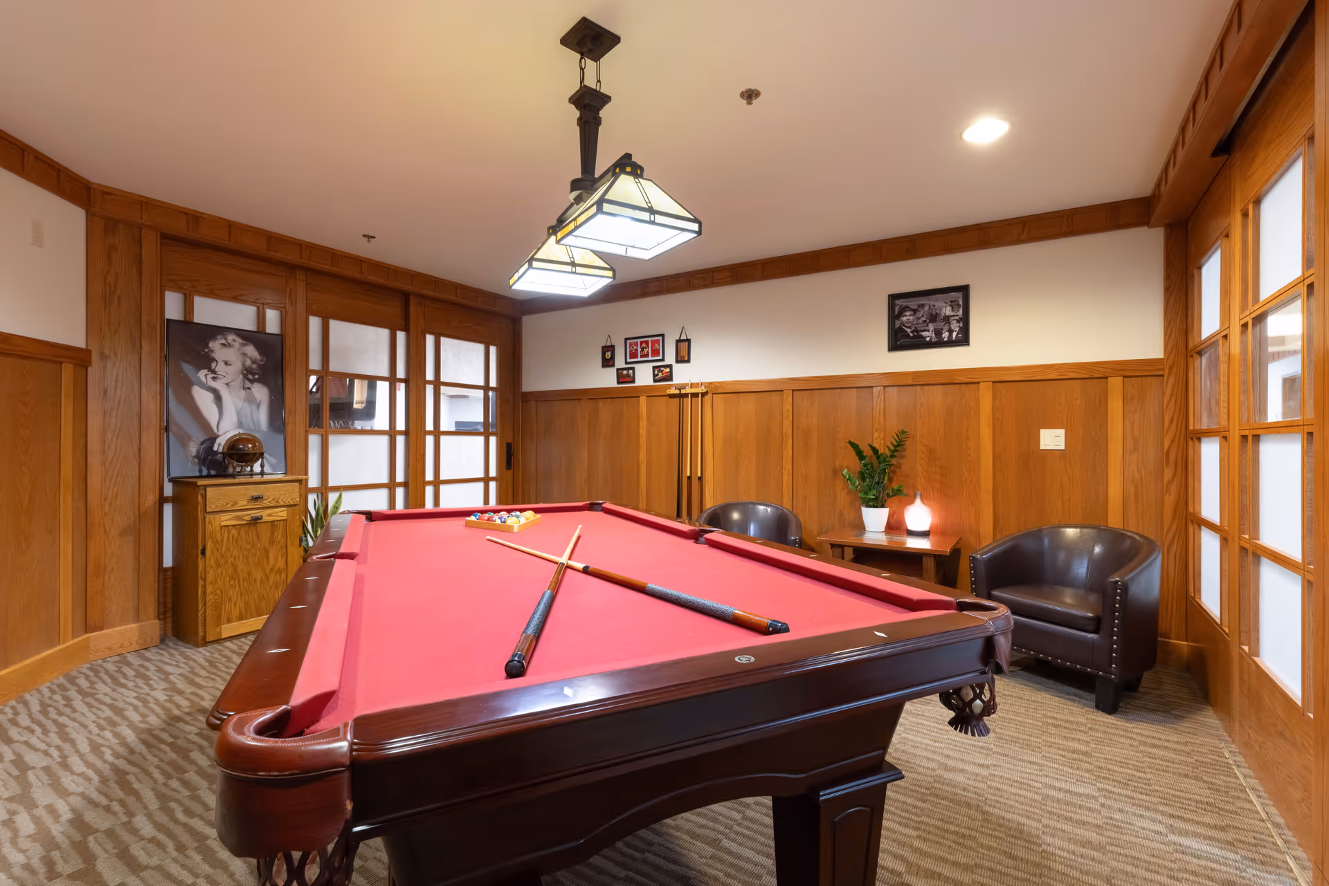 A cozy game room with a red felt pool table in the center, two pool cues resting on the table, and a rack of billiard balls. The room features wood-paneled walls, a carpeted floor, two black leather chairs, a small wooden side table with a plant and a decorative light, and framed black-and-white photographs on the walls. There is a wooden cabinet with a vintage radio and a black-and-white portrait of Marilyn Monroe. The room is softly lit by a ceiling light fixture.