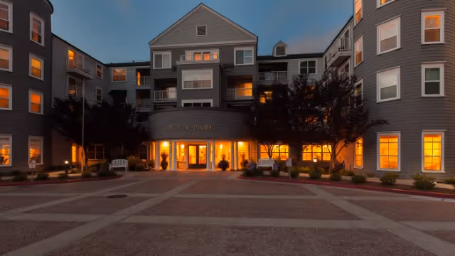 Front entrance of the Byron Park senior living building at dusk with illuminated windows and a paved driveway.
