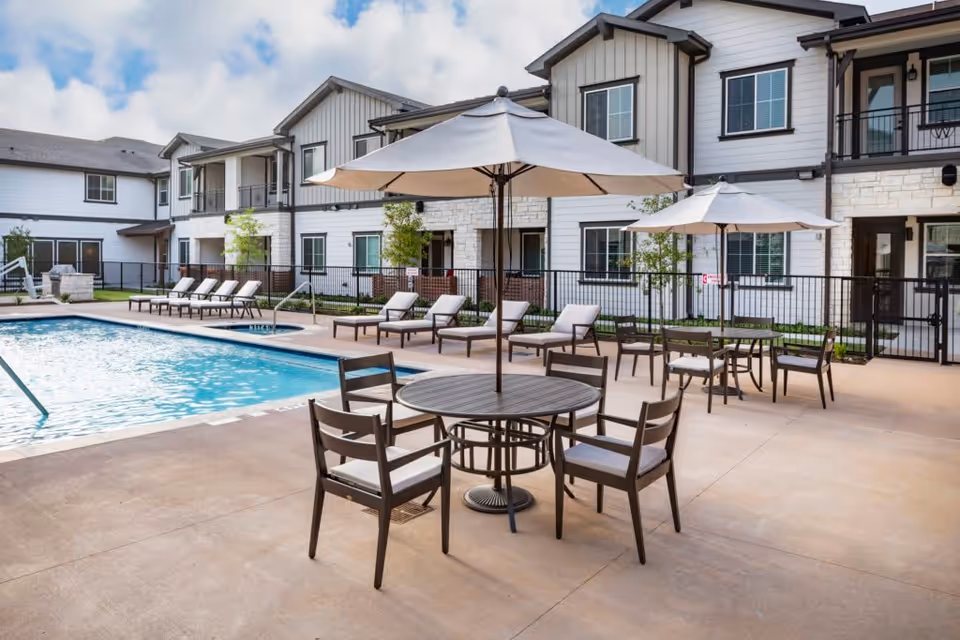 Outdoor pool area at The Preserve at Spring Creek with lounge chairs lined up along the poolside and round tables with umbrellas and chairs on the patio. The background shows a two-story building with white siding and stone accents under a partly cloudy sky.