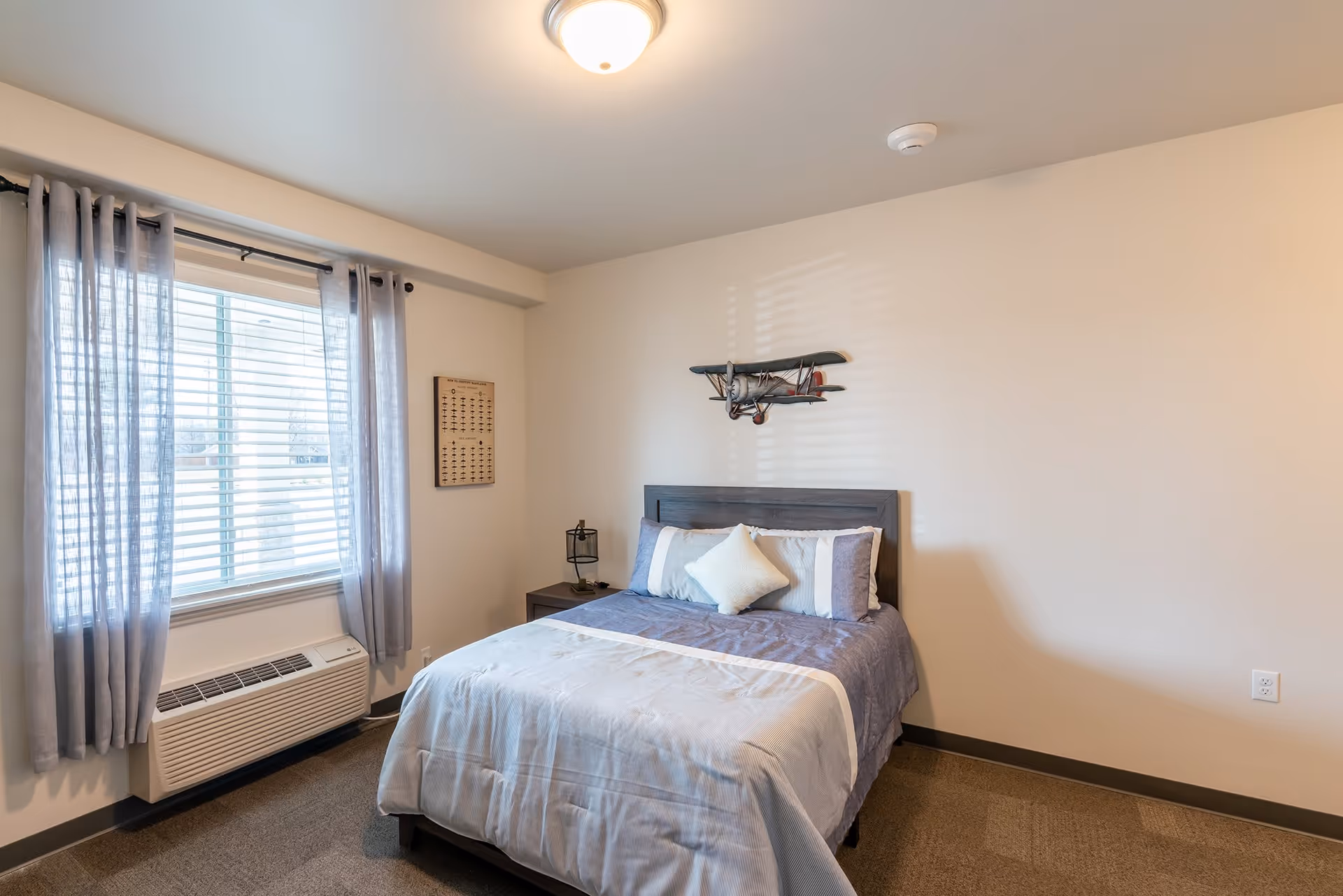 A neatly made bed with blue and white bedding in a simple bedroom. There is a window with sheer gray curtains and blinds on the left wall, a nightstand with a lamp next to the bed, and a decorative airplane mounted on the wall above the headboard. The room has beige walls and carpeted floor.