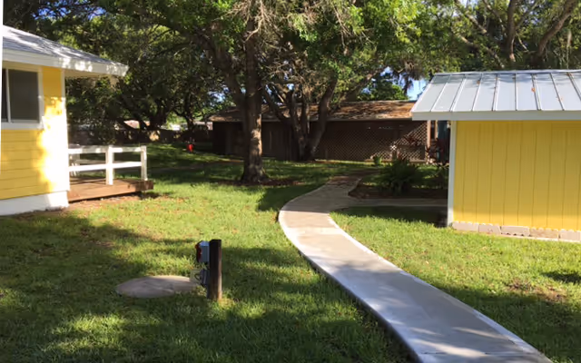 A sunny outdoor area with a curved concrete pathway leading between two yellow buildings. There are trees providing shade and a grassy lawn surrounding the path.
