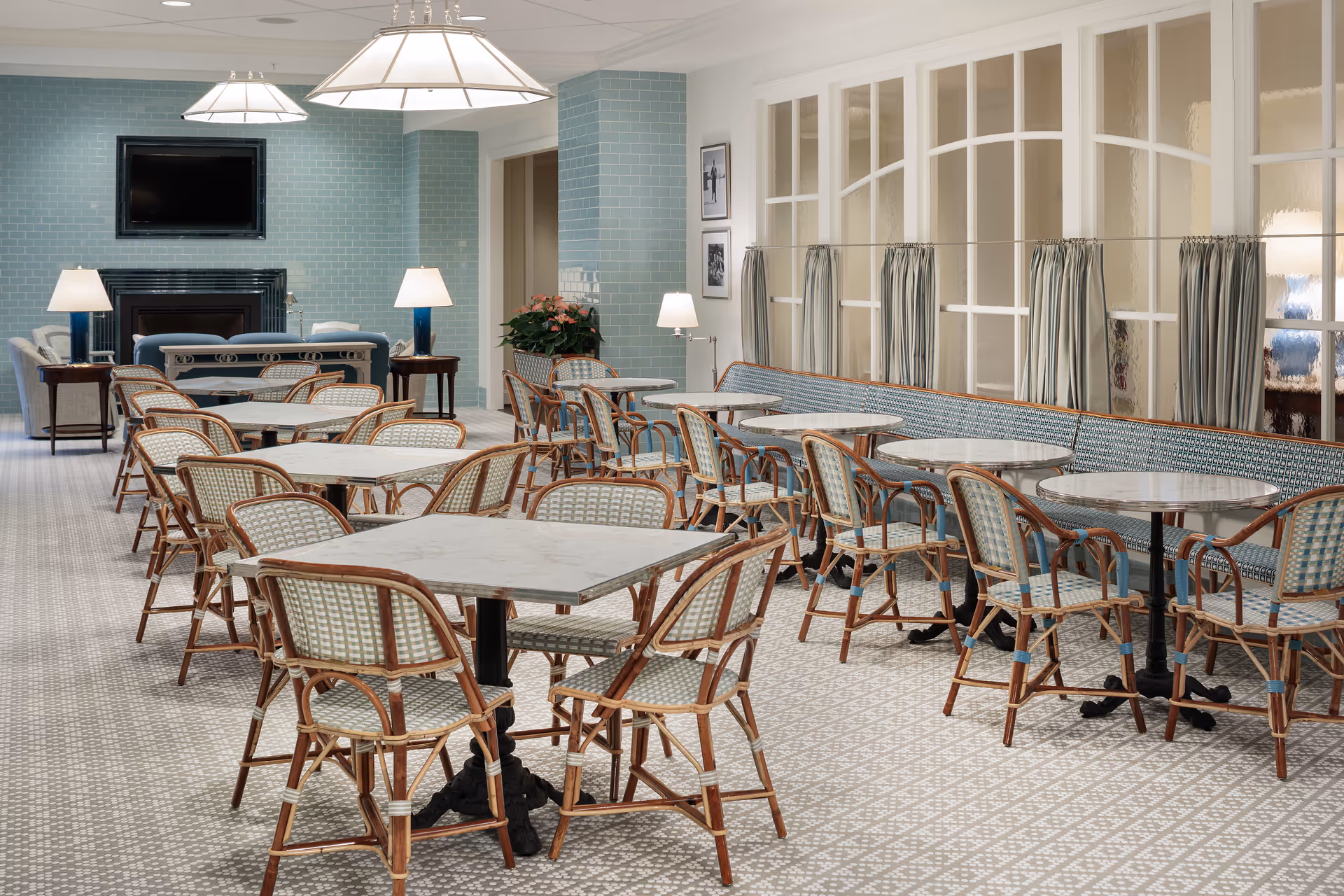 Bright dining room with multiple marble-top tables and wicker chairs arranged around a long banquette and lamps.
