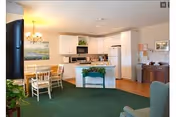 A bright and spacious assisted living facility common area featuring a kitchen with white cabinets and appliances, a dining table with chairs, a green carpeted floor, and a chandelier. There is a small green table with a plant on it and a wooden sideboard with framed pictures on the wall.