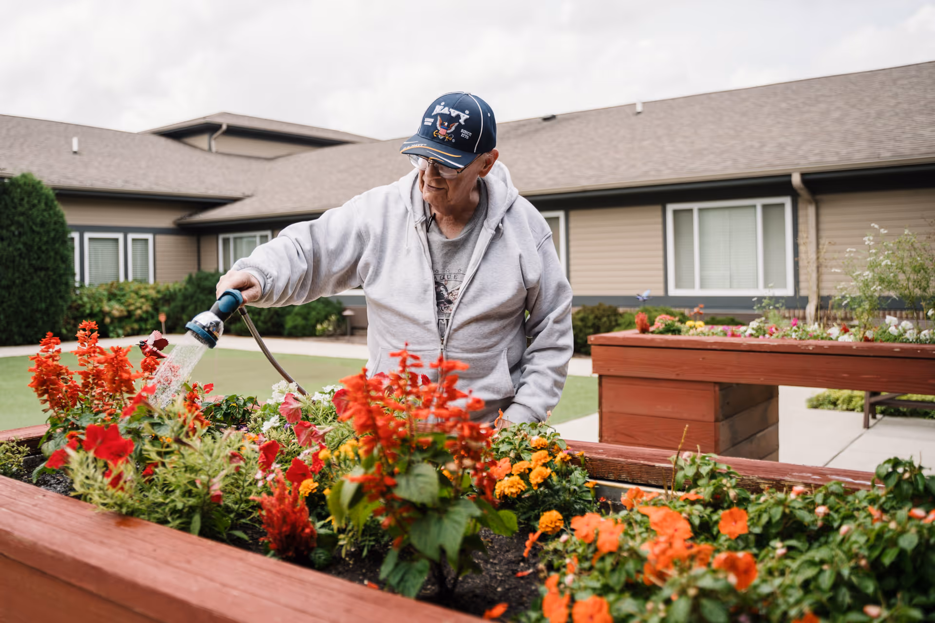 An elderly man wearing a navy cap and gray hoodie waters colorful flowers in a raised garden bed outside a single-story building under a cloudy sky.