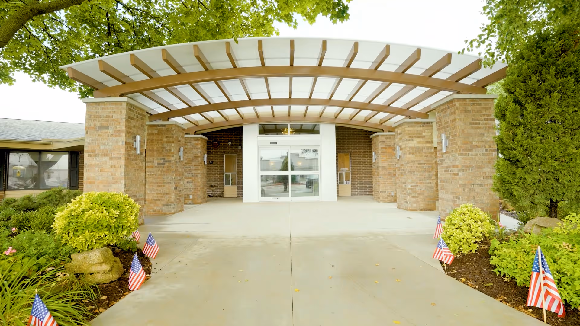 Covered brick entrance with a curved canopy, glass double doors, landscaped beds and small American flags along the walkway.