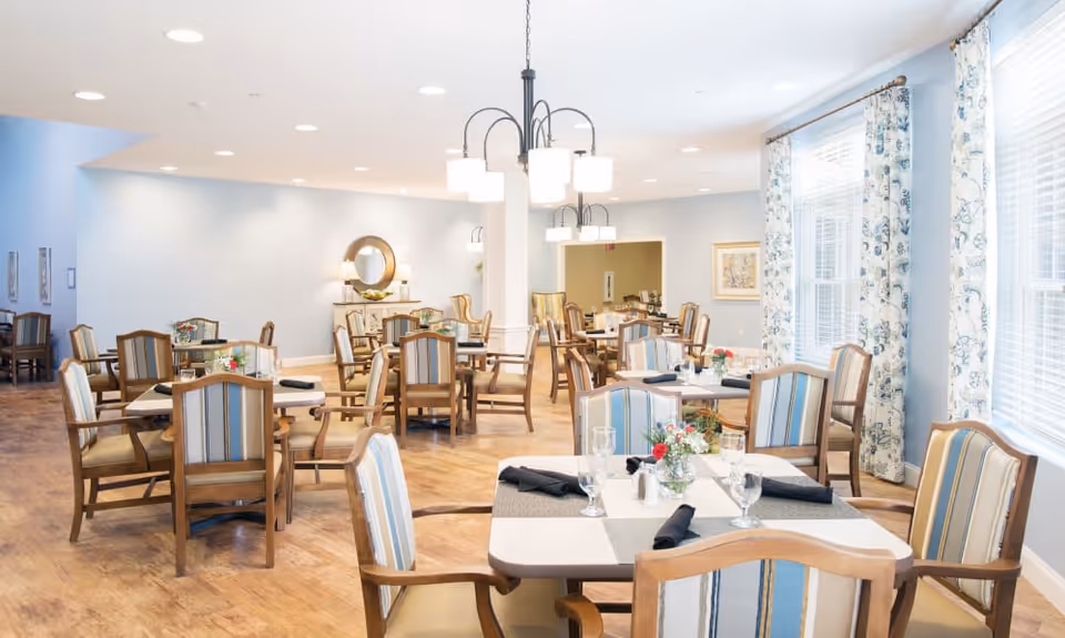 A bright and spacious dining room with multiple wooden tables and chairs arranged neatly. Each table is set with glasses, black napkins, and small floral centerpieces. The room features light blue walls, large windows with floral curtains, and ceiling lights providing ample illumination.