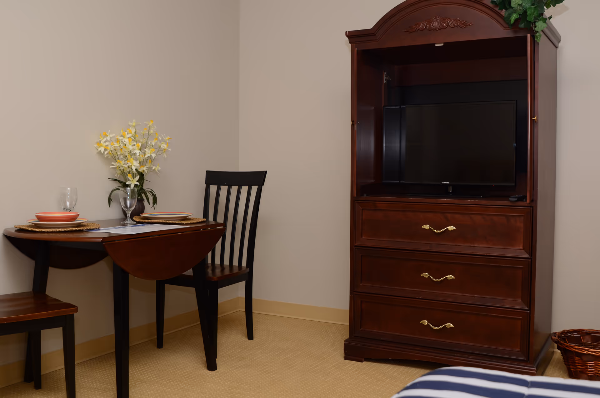 A small dining area with a dark wooden table set for two, including plates, glasses, and a vase with yellow and white flowers. Next to the table is a dark wooden chair. To the right, there is a dark wooden entertainment cabinet with a flat-screen TV inside and three drawers below. The room has beige walls and carpeted flooring.