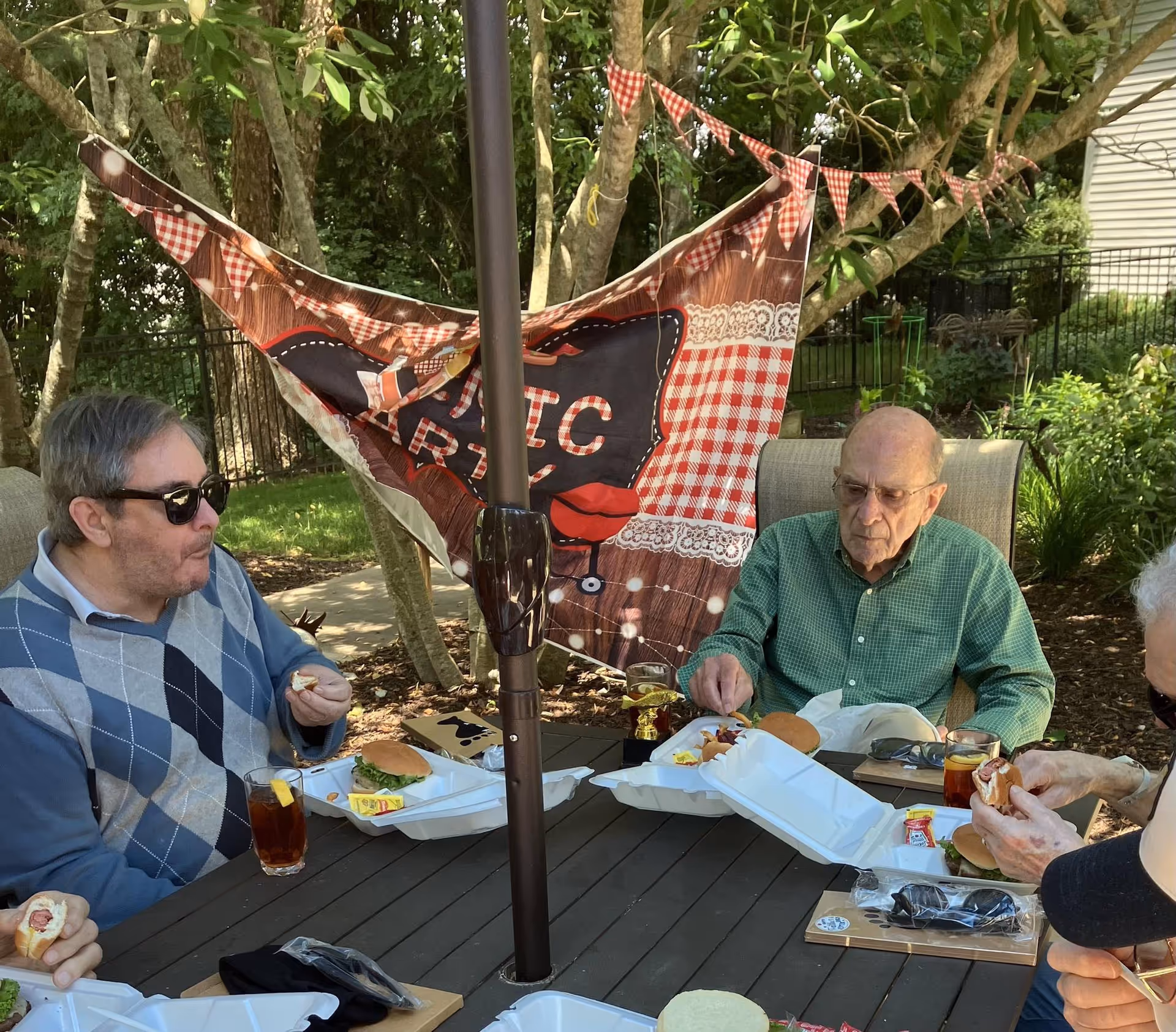 People sitting around an outdoor patio table eating meals from takeout containers under a decorative picnic banner.
