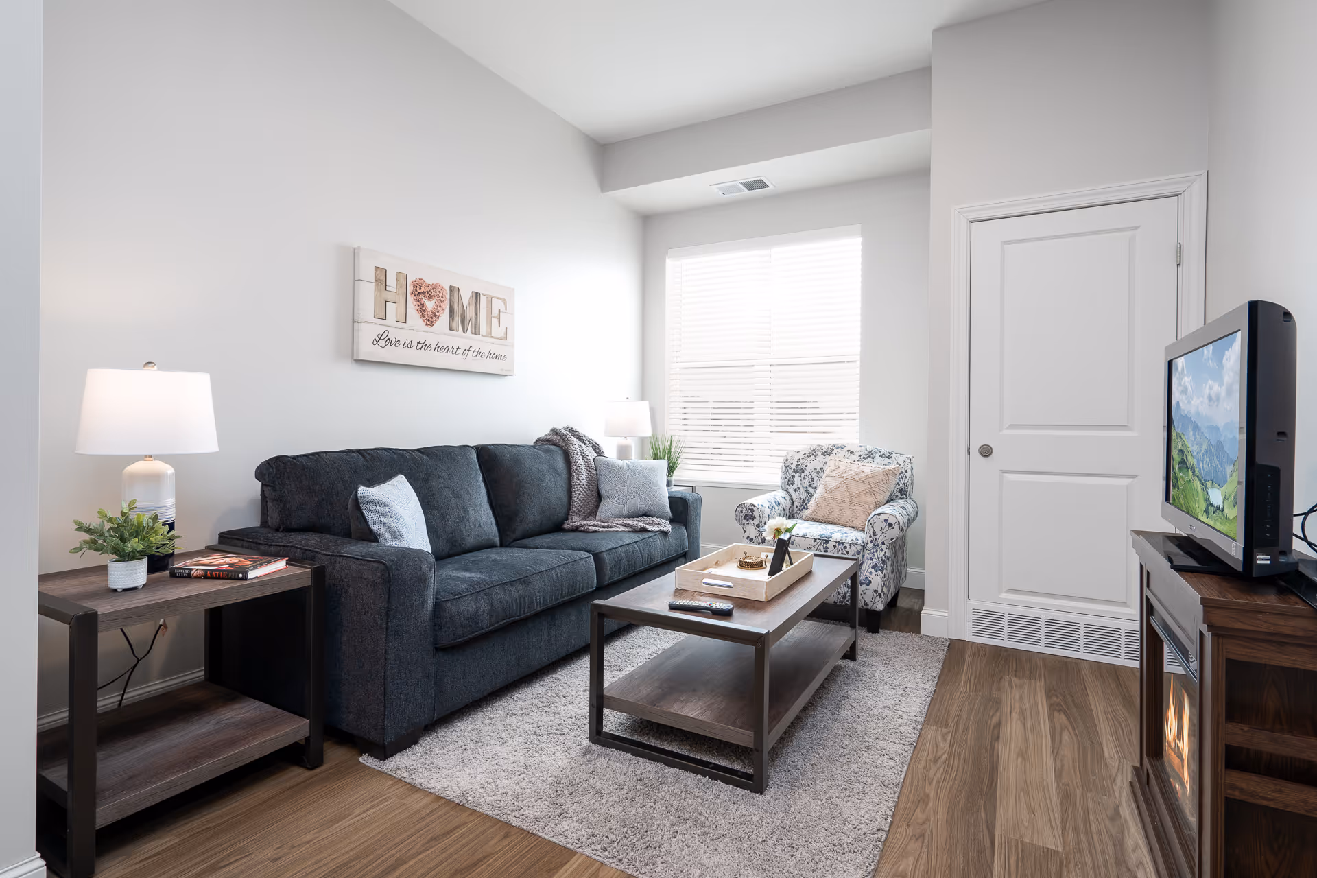 A cozy living room with a dark gray sofa adorned with light blue and gray pillows, a floral armchair, and a wooden coffee table with a tray and remote controls. There is a side table with a lamp and a small plant, a window with blinds letting in natural light, and a TV on a wooden stand with an electric fireplace below. A wall decoration above the sofa reads 'HOME Love is the heart of the home.'