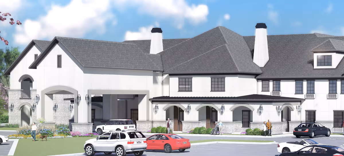 Exterior view of a large senior living facility building with white walls and a gray roof under a blue sky with some clouds. Several cars are parked in front, and a few people are walking or standing near the entrance, including one person in a wheelchair. There are some landscaped plants and flowers near the building.