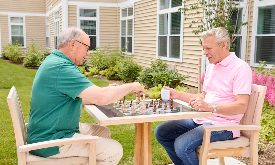 Two elderly men sitting outside at a wooden table playing chess. One man is wearing a green shirt and glasses, and the other is wearing a pink polo shirt holding a white mug. They are in a garden area next to a beige building with windows and green shrubs.
