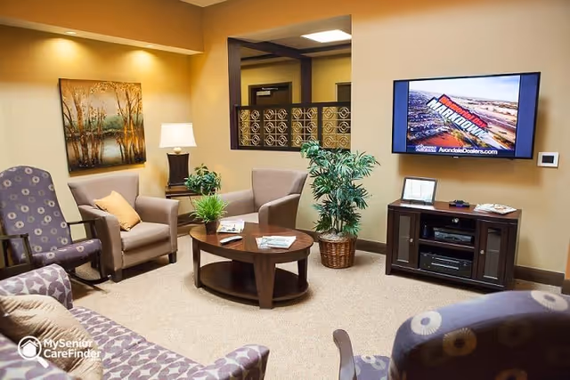 A cozy living room area with beige walls and carpeted floor, featuring a patterned sofa and armchairs arranged around a wooden coffee table with magazines and a small plant. A flat-screen TV is mounted on the wall above a dark wooden media console, and a large potted plant is placed next to the console. A framed painting and a table lamp are visible on a side table in the corner.