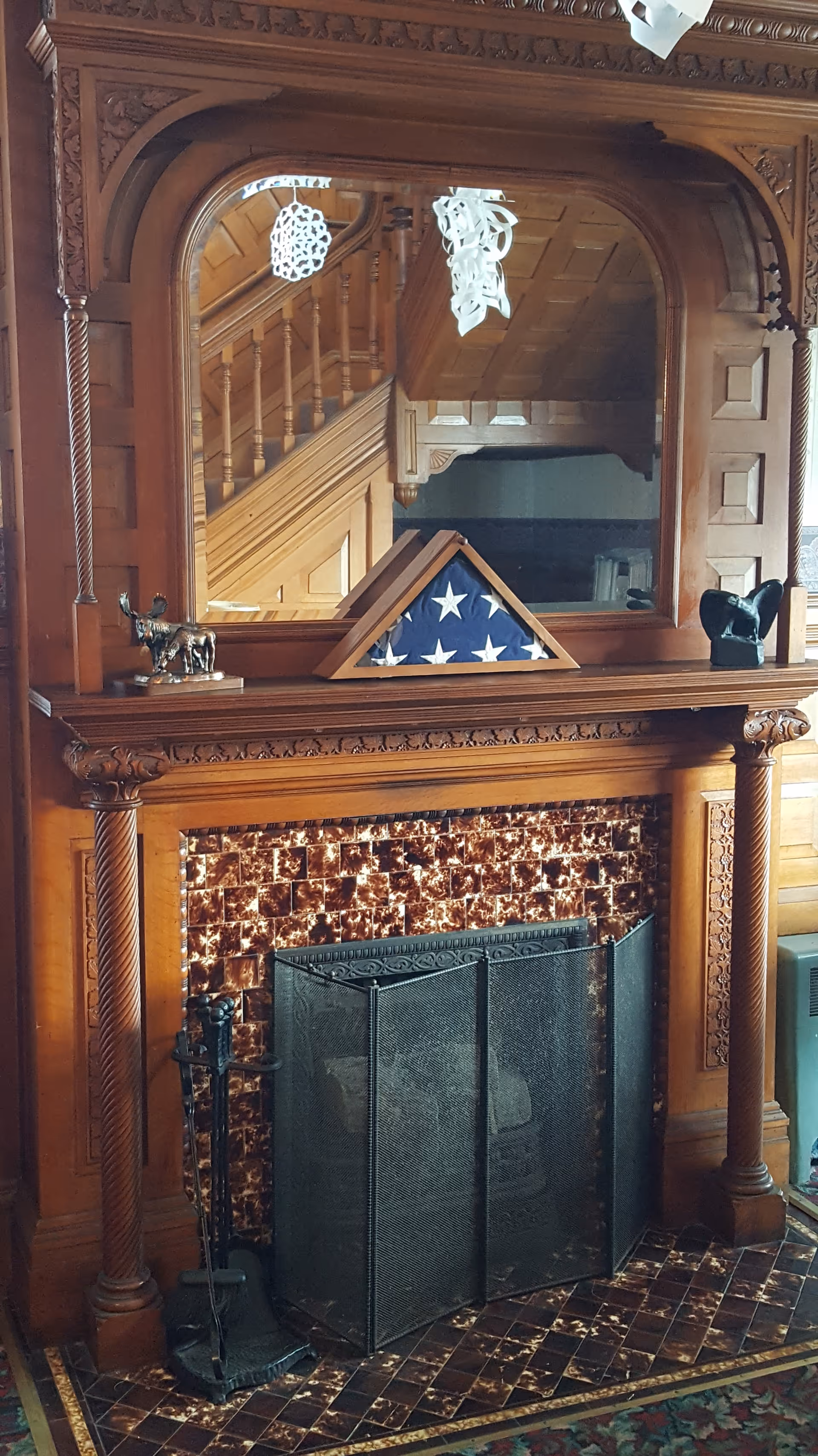 Ornate wooden fireplace mantel with a mirrored overmantel, a folded American flag on the mantel, and a tiled hearth with a fireplace screen.