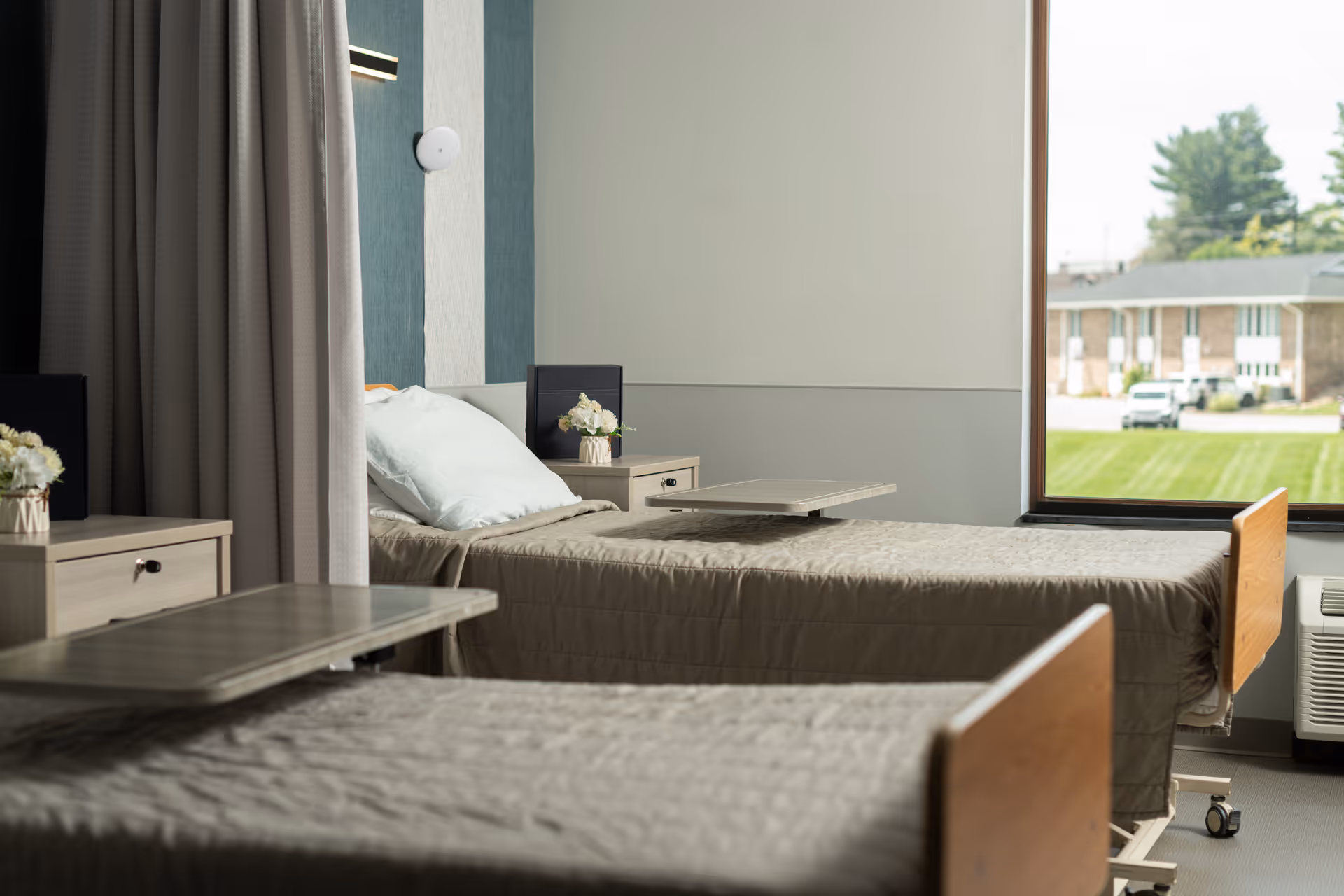 A bright and clean room with two hospital-style beds, each with a beige quilted blanket and a pillow. Each bed has an adjustable overbed table. There are two small bedside tables with flower arrangements. A large window shows a view of a green lawn and buildings outside. The room has light-colored walls and a curtain partially drawn between the beds.