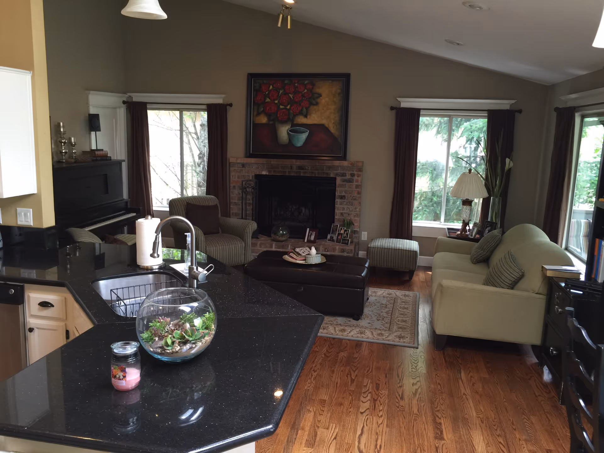 Open-plan living room and kitchen with a black countertop island, sofa and armchairs facing a brick fireplace and large windows.
