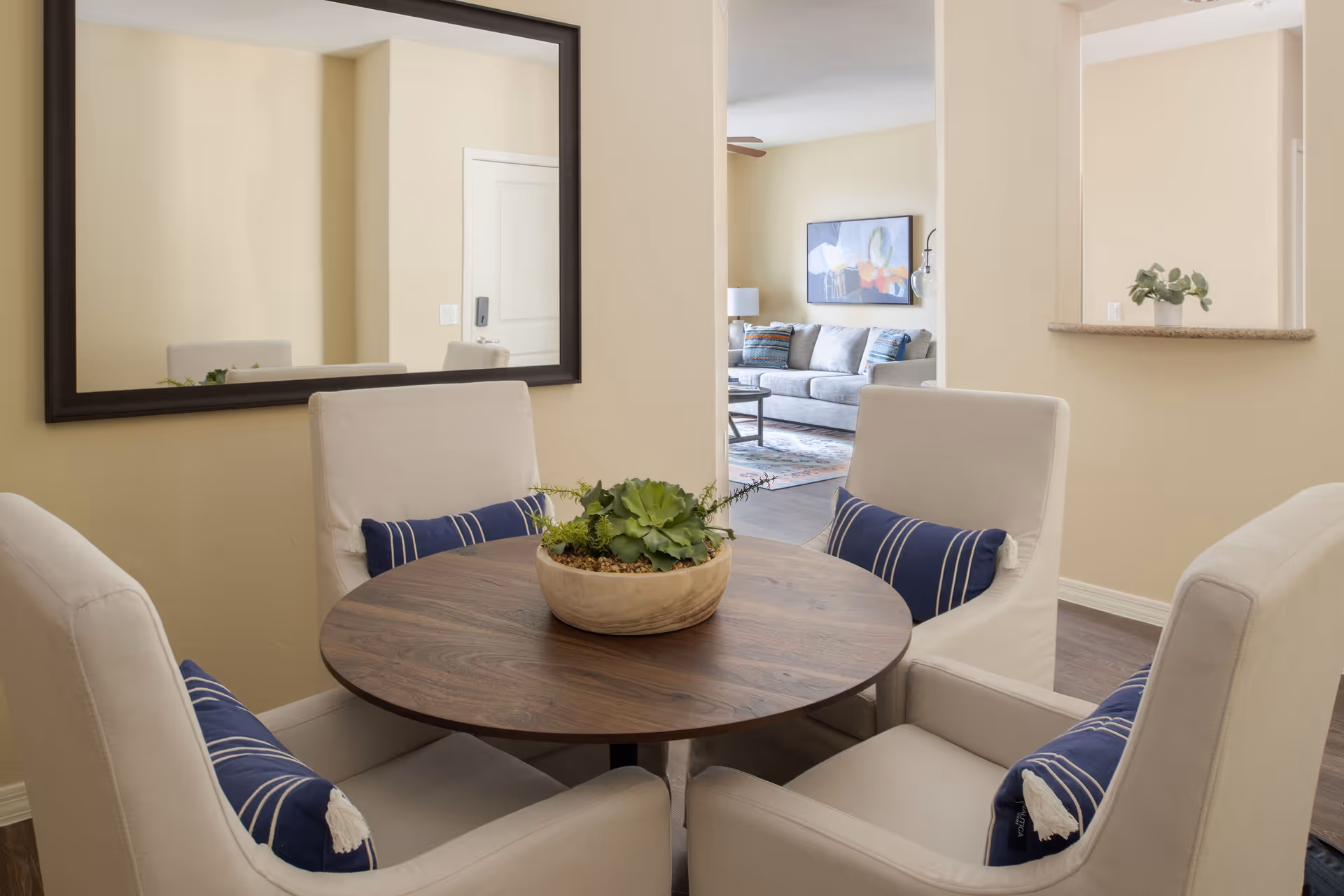A cozy dining area with a round wooden table surrounded by four beige upholstered chairs, each with a blue and white striped pillow. A large rectangular mirror hangs on the wall, reflecting part of the room. In the background, a living room with a gray sofa, a coffee table, a lamp, and wall art is visible through an open doorway.