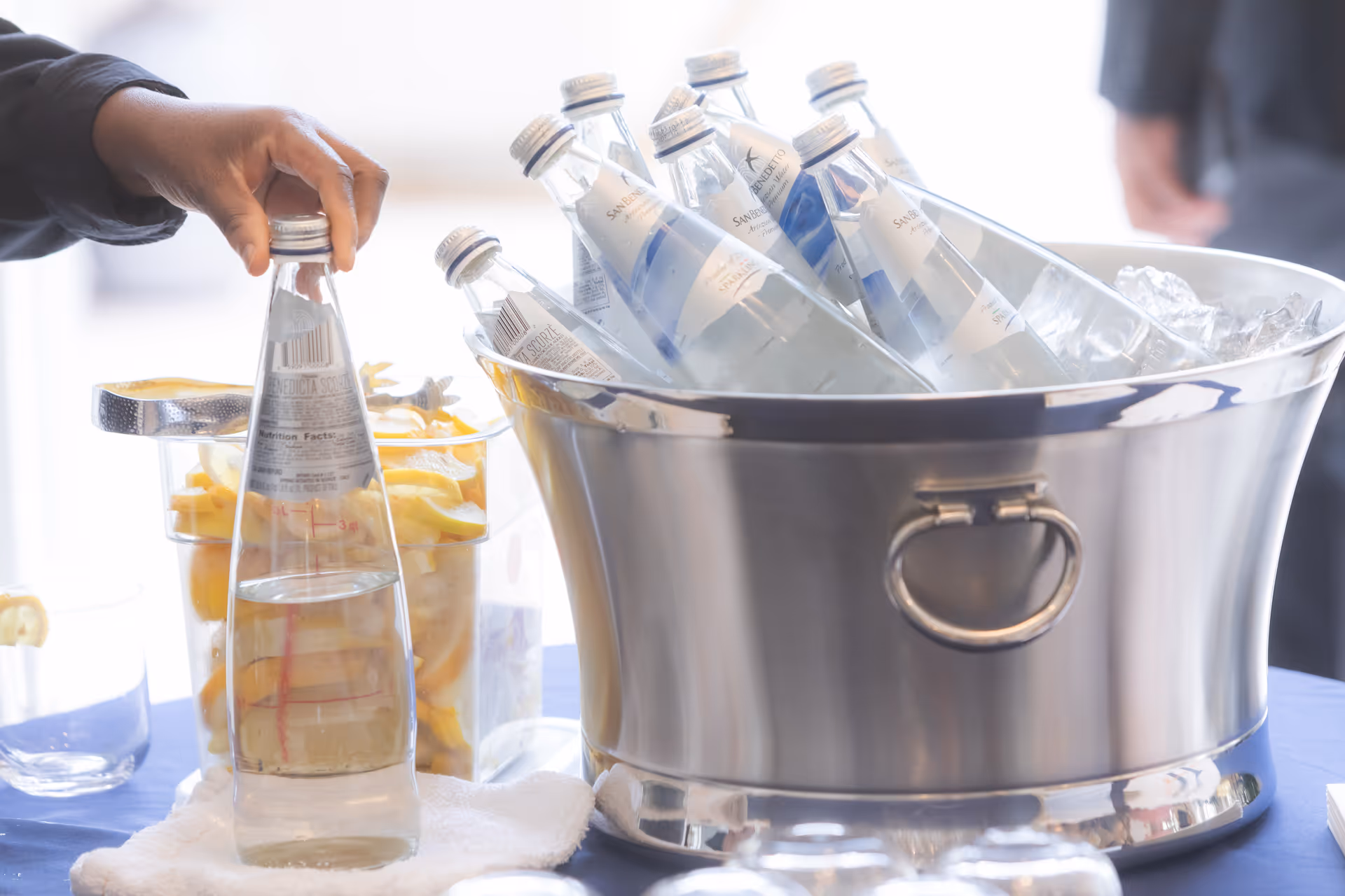 A hand reaching for a chilled bottled water from an ice-filled metal bucket beside a container of lemon slices.