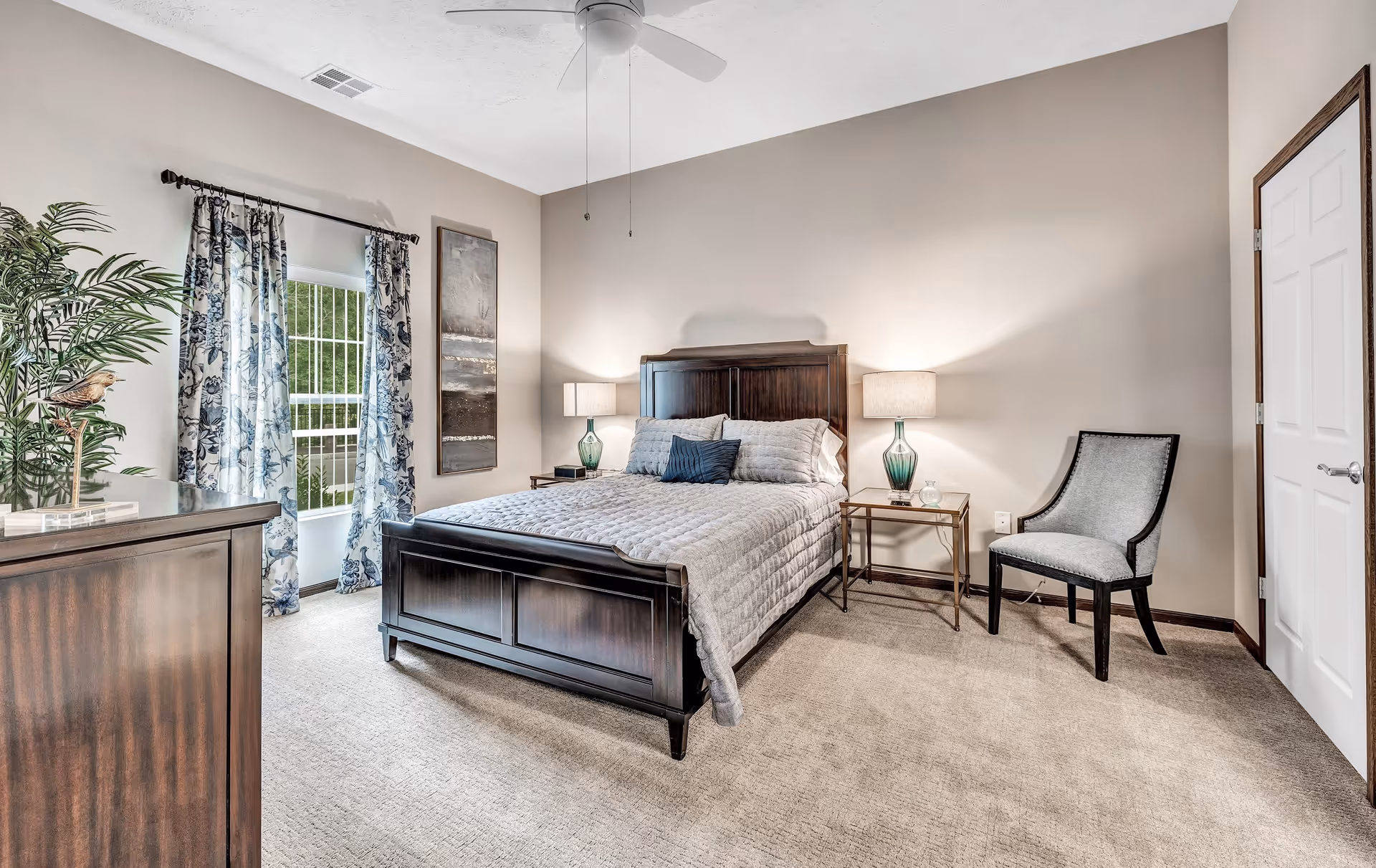 A neatly arranged bedroom with a dark wooden bed frame, a gray quilted bedspread, and three pillows. There are two matching bedside tables with green glass lamps on each side of the bed. A gray upholstered chair is placed near a white door. A dresser with a decorative bird sculpture and a tall green plant is on the left side. The room has beige walls, carpeted floor, a ceiling fan, and a window with floral curtains letting in natural light.