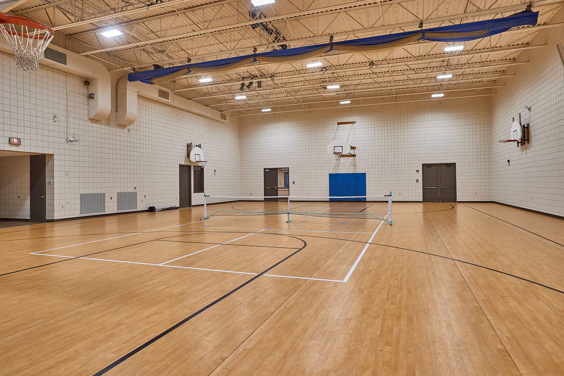 Indoor gymnasium with wooden flooring, multiple basketball hoops mounted on the walls, and a net set up in the middle of the court. The walls are light-colored with ventilation ducts near the ceiling and several doors along the walls.