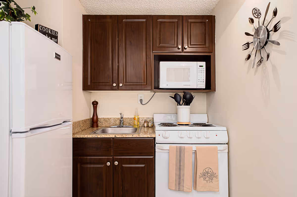 A compact kitchen area featuring a white refrigerator on the left, dark wooden cabinets above and below a granite countertop with a small sink. A white microwave is mounted above a white stove with two beige towels hanging on the oven door. Kitchen utensils are stored in a white container on the stove. A decorative clock with utensils as clock hands is mounted on the wall to the right.