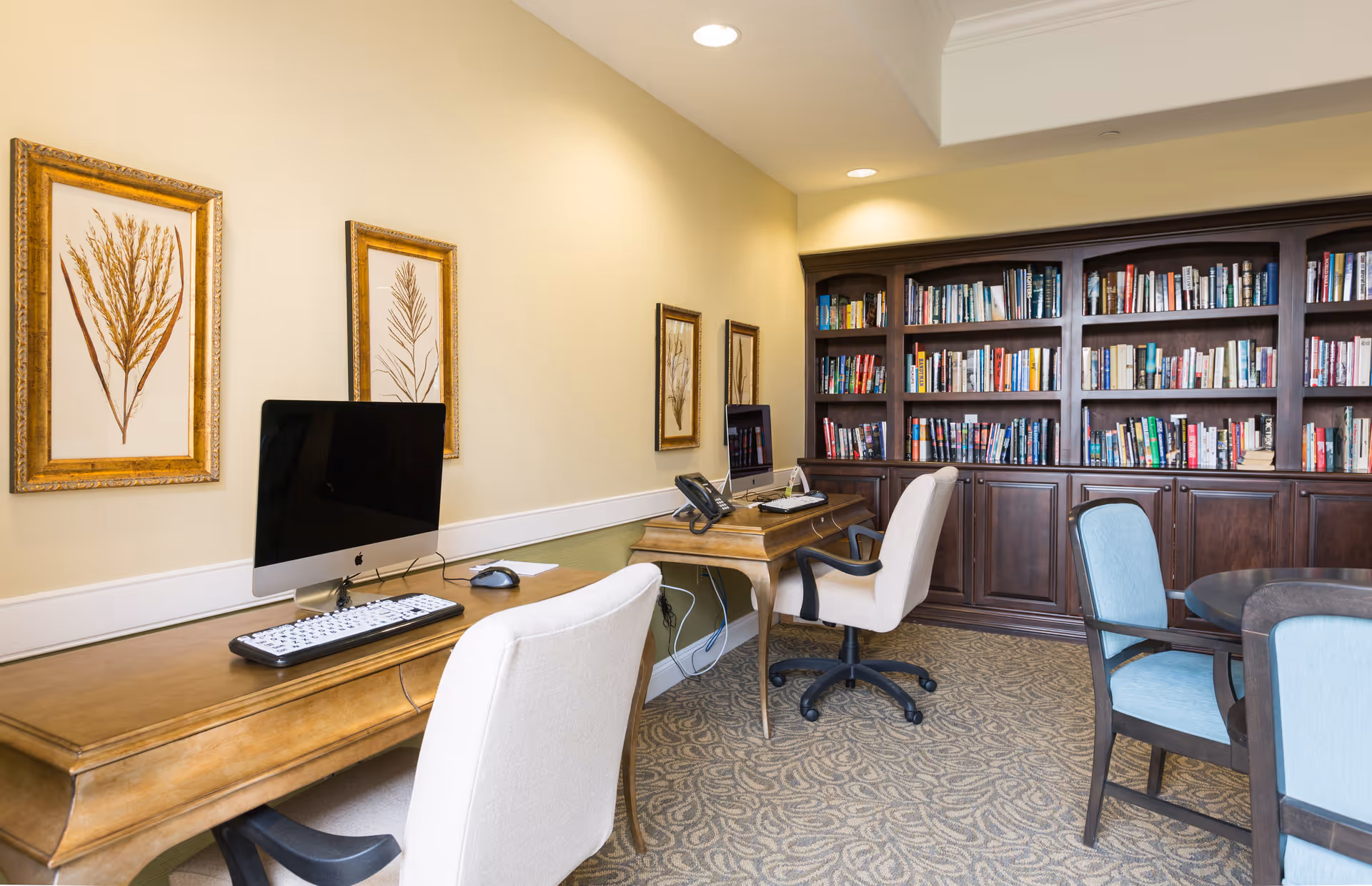 A cozy room with two wooden desks each equipped with a computer and office chair. The room features a large dark wood bookshelf filled with books along the back wall. There are framed botanical prints on the light yellow walls and a round table with blue upholstered chairs in the foreground.
