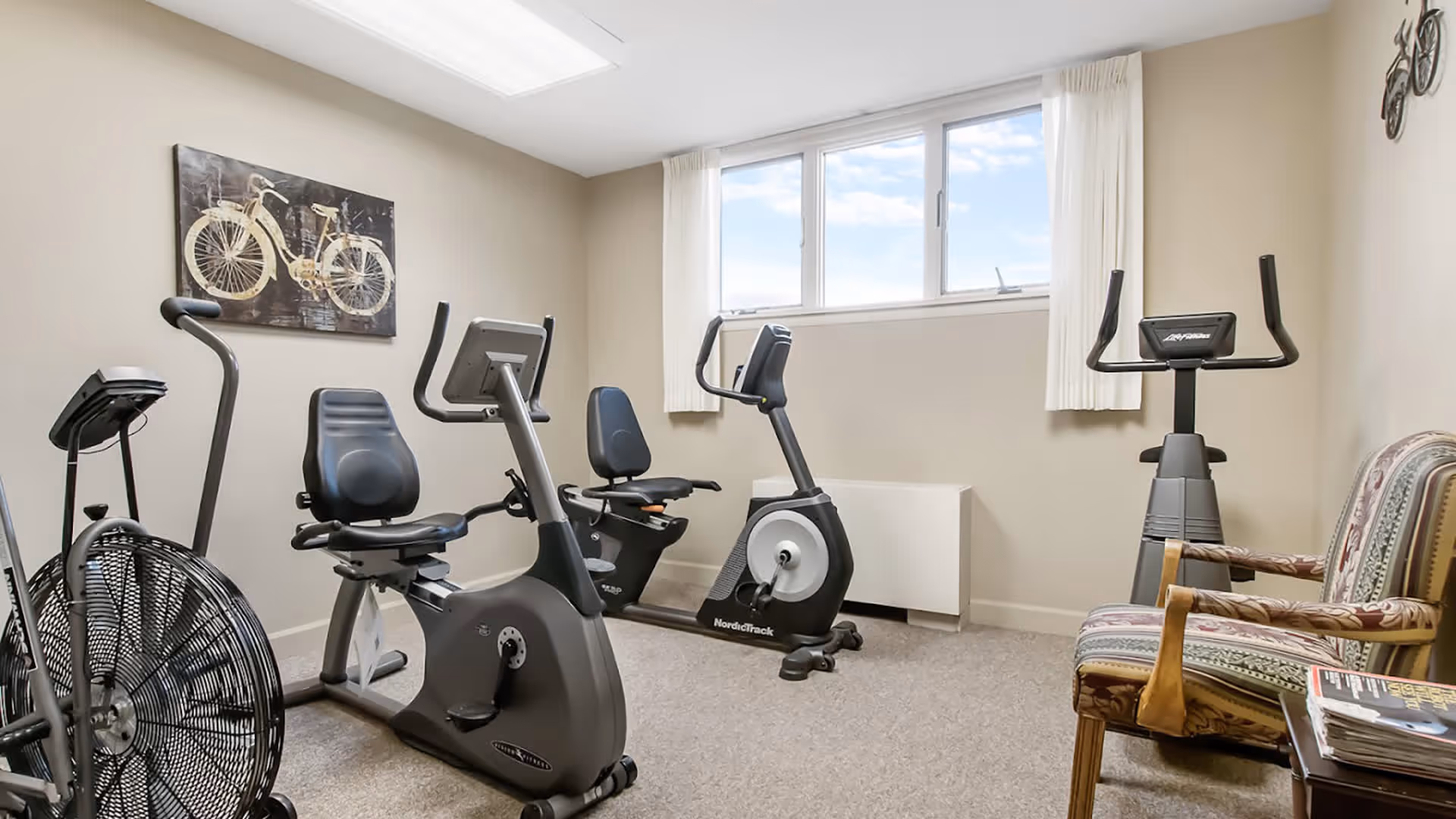 A small exercise room with three stationary bikes, a fan bike, a window with white curtains, a painting of a bicycle on the wall, and two patterned armchairs next to a small table with magazines.