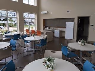 Interior view of a senior living facility dining area with round tables and chairs arranged neatly. Each table has a small floral centerpiece. Large windows allow natural light to fill the room, and a kitchen area with appliances is visible in the background.