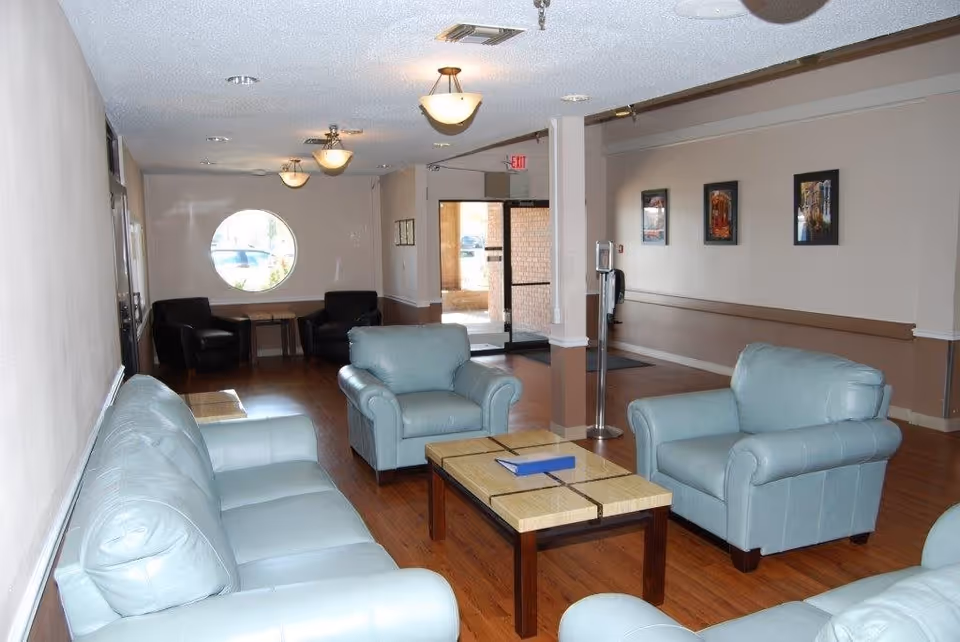 A seating area in a senior living facility with light blue leather sofas and armchairs arranged around a wooden coffee table. The room has wooden flooring, beige walls with a brown trim, three ceiling light fixtures, framed pictures on the wall, and a circular window near the entrance door.