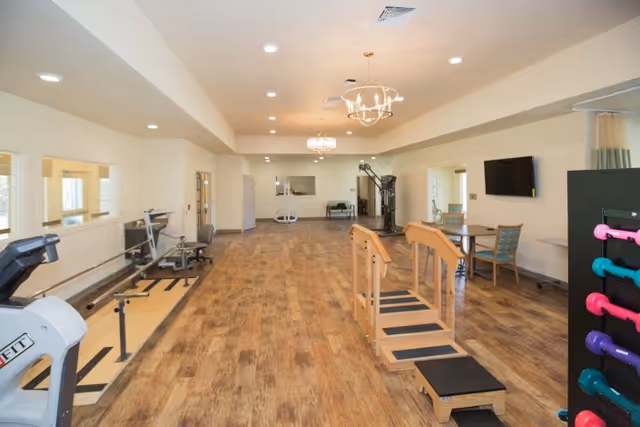 A spacious rehabilitation and exercise room with wooden flooring, various exercise equipment including a treadmill, parallel bars for walking practice, weight machines, and a rack of colorful dumbbells. The room is well-lit with ceiling lights and has a wall-mounted TV and seating area with chairs and a table.