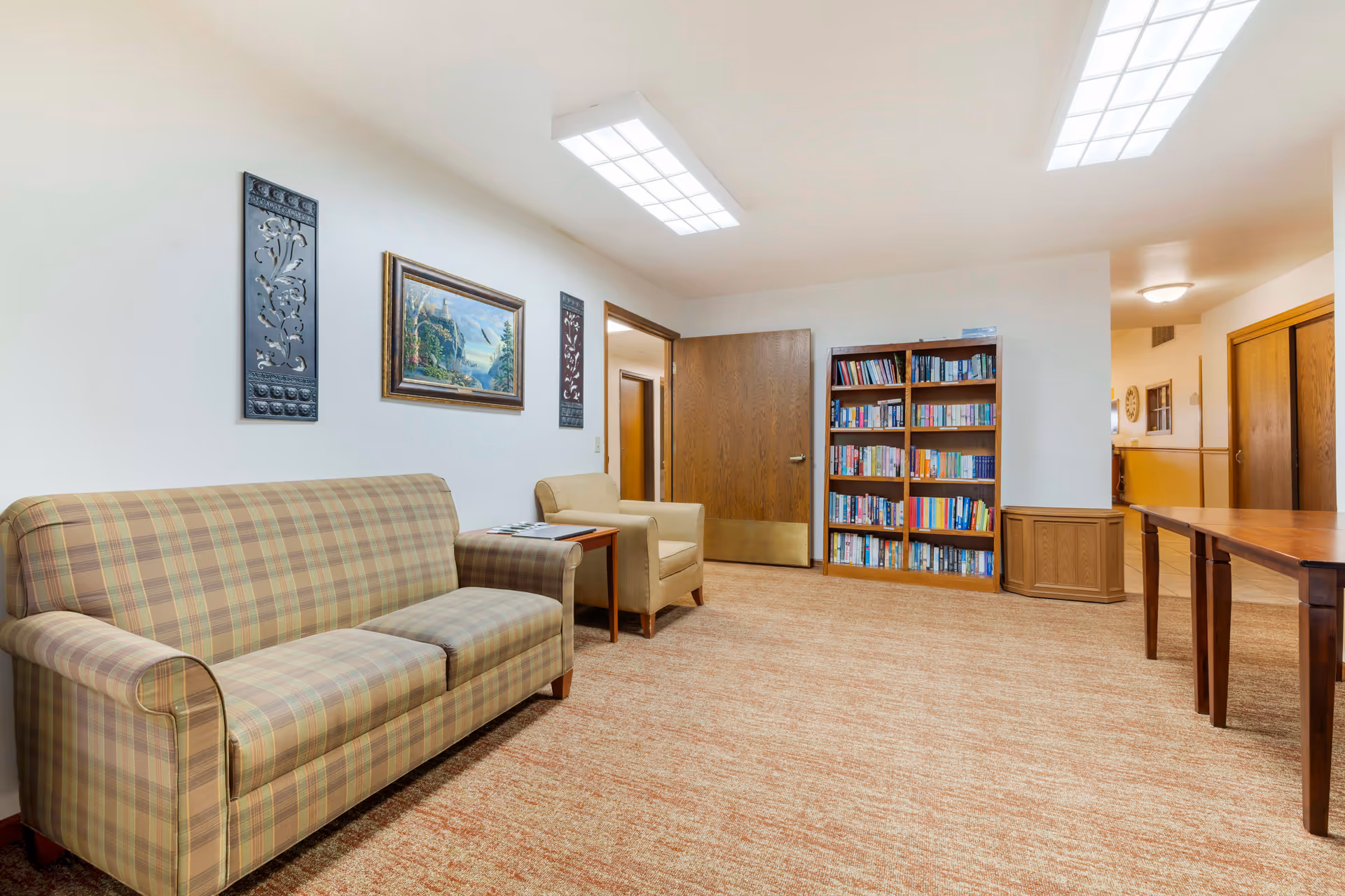 A cozy room with a plaid sofa and a beige armchair next to a small wooden side table. On the wall above the sofa are two decorative black panels and a framed painting of a landscape. Across the room is a wooden bookshelf filled with books, and a wooden table is partially visible on the right. The room has beige carpet and white walls with wooden doors and trim.