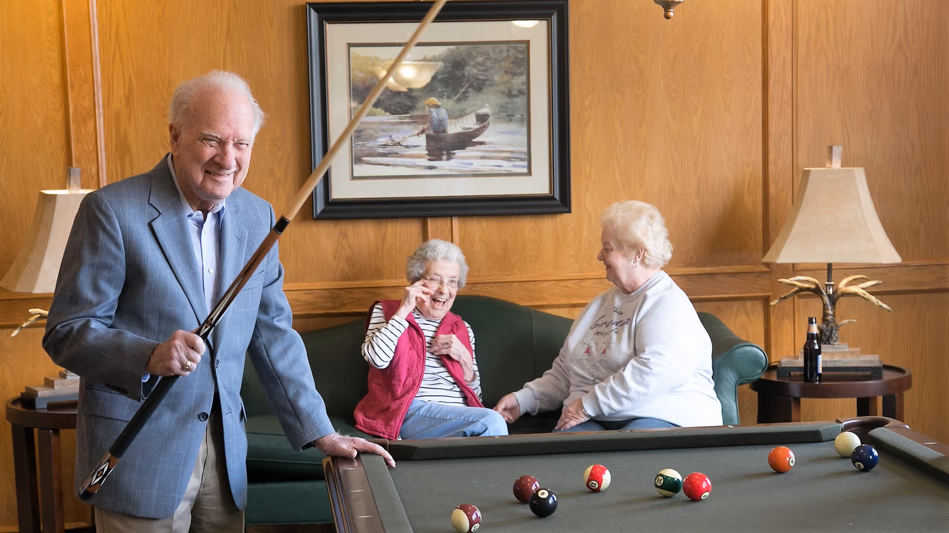 An elderly man holding a pool cue stands next to a pool table, smiling. Two elderly women sit on a green couch behind the pool table, engaged in conversation and laughing. The room has wood-paneled walls, a framed painting of a person in a canoe, and two table lamps on side tables.