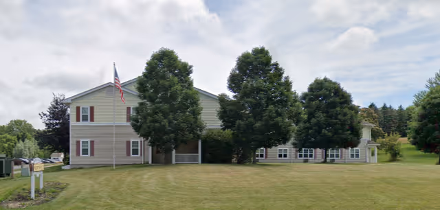 Two-story beige residential building set behind three large trees with an American flag and a mowed front lawn.
