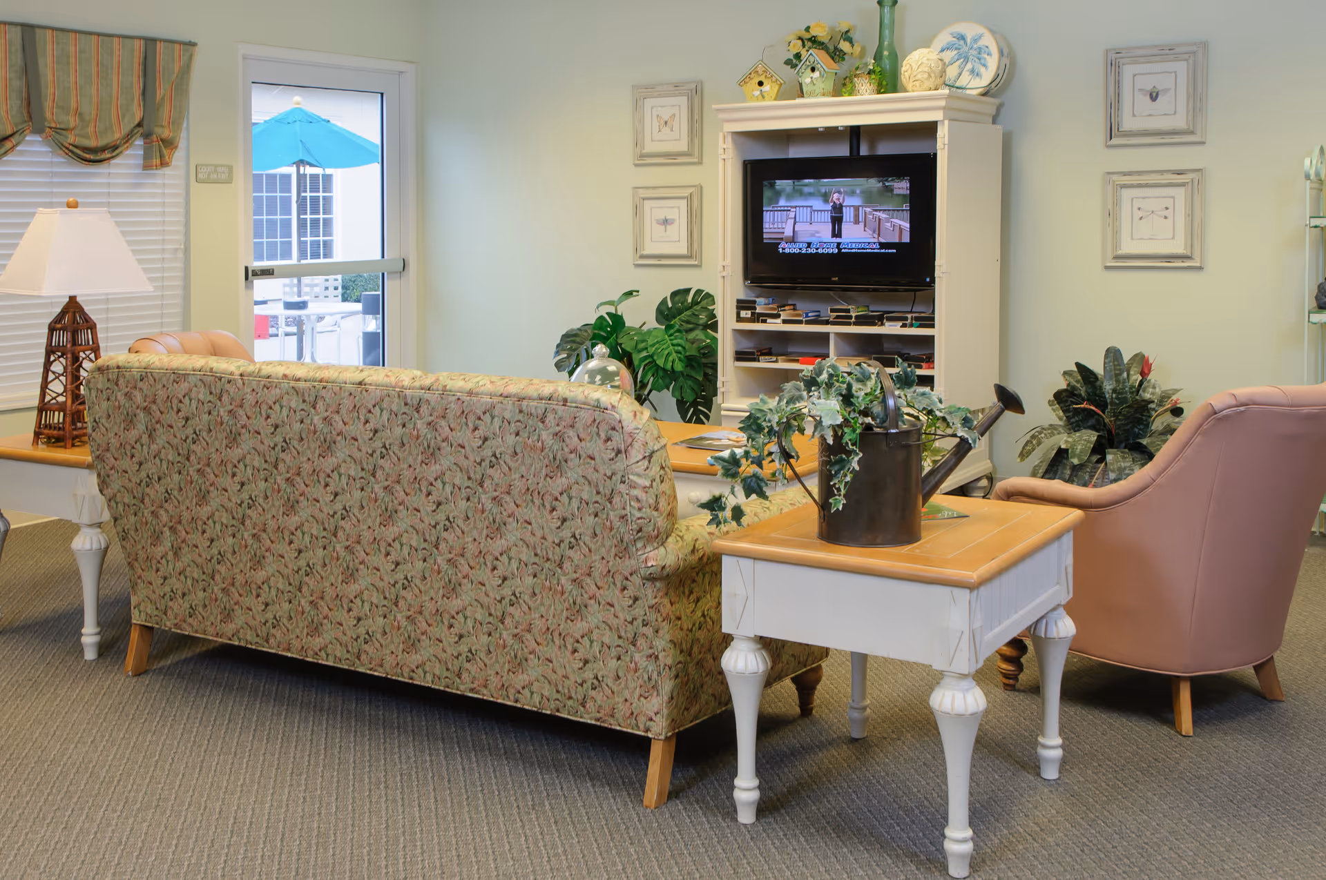 A cozy living room area with a floral patterned sofa, a pink armchair, and a white wooden side table with a watering can and green plant on it. A white entertainment center holds a television playing a program. The room has light-colored walls decorated with framed pictures and several potted plants. A door with a window shows an outdoor patio with a blue umbrella.