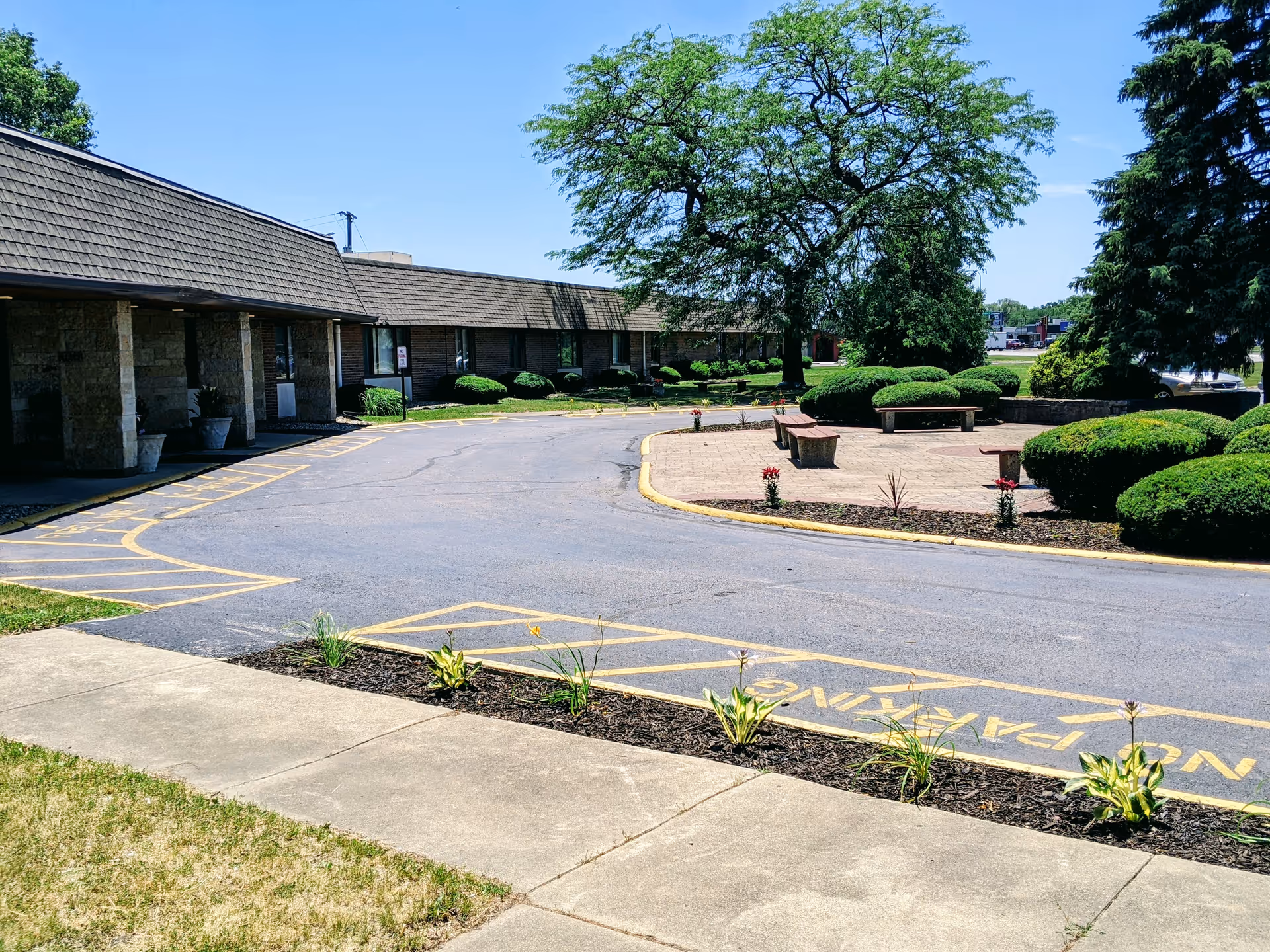 Front exterior of a single-story senior living building with a circular driveway, landscaped beds, and benches beneath trees.
