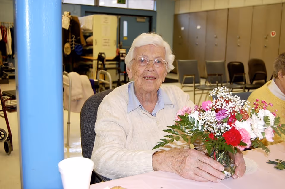 An elderly woman with white hair and glasses sitting at a table indoors, holding a vase with a colorful bouquet of flowers. She is wearing a light-colored sweater over a blue collared shirt. In the background, there are chairs, lockers, and other items indicating a communal or activity room.