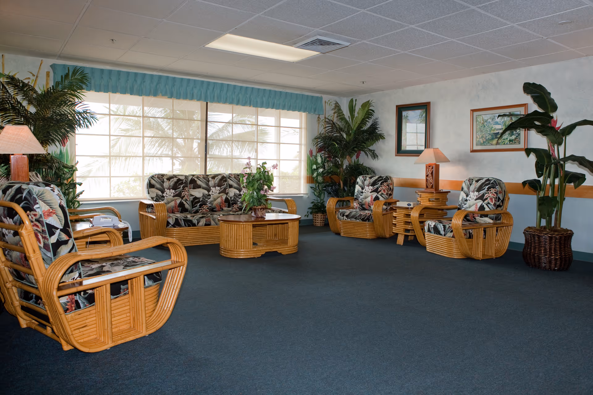 A spacious living room area with wicker furniture including a sofa, armchairs, and a coffee table. The cushions have a floral pattern. There are several potted plants around the room, two table lamps on side tables, and framed artwork on the walls. Large windows with blinds and a blue valance let in natural light.