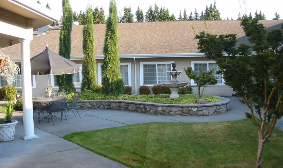 Outdoor courtyard with a central stone planter and fountain, patio table with umbrella, and the facility building with windows in the background.