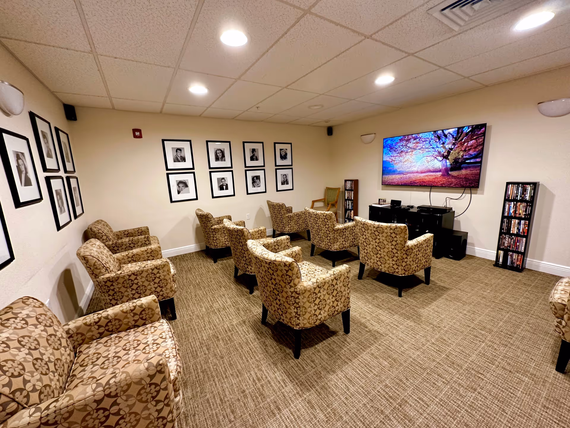 A small media/lounge room with patterned armchairs arranged in rows facing a large wall-mounted TV and framed black-and-white photos on the walls.