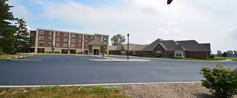 Exterior view of a senior living facility with a large paved driveway and parking area in front. The building has multiple sections with varying roof heights, including a three-story section on the left and a single-story section on the right. There are some trees and greenery around the building under a partly cloudy sky.