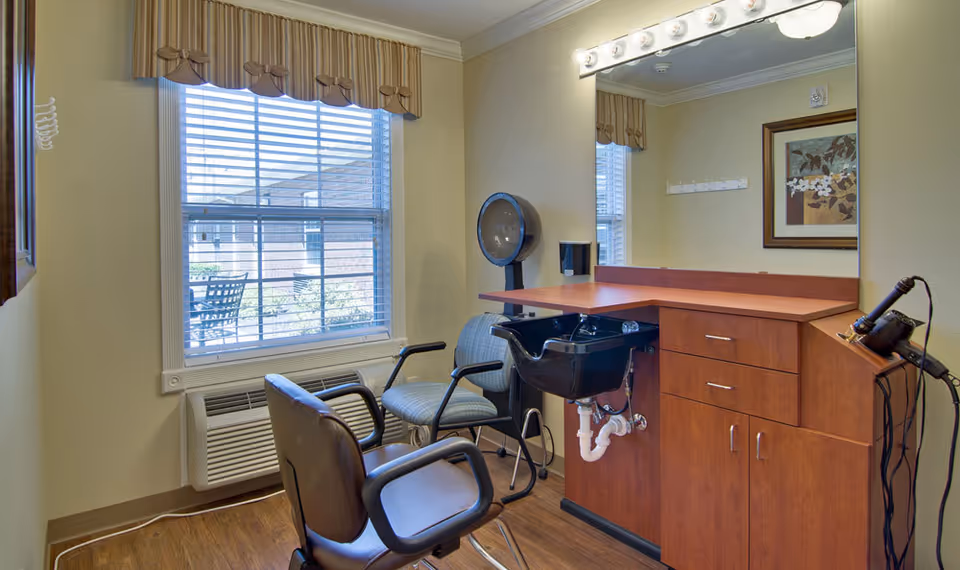 Interior of a senior living facility hair salon with a styling chair, a washing station with a black sink, a large mirror with lights above it, a window with blinds and a valance, and a hair dryer on the counter.