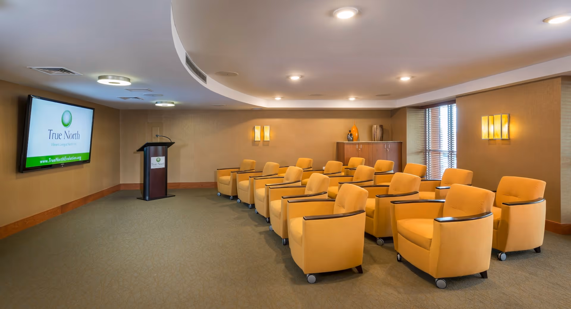 A small conference or presentation room with two rows of yellow armchairs on wheels facing a podium and a large wall-mounted screen displaying the True North logo and website. The room has beige walls, carpeted floor, ceiling lights, and a window with blinds on the right side.